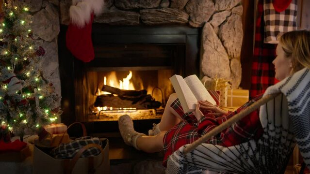Cinematic shot of young woman reading book and relaxing at cozy burning fireplace at home decorated for Christmas. Relaxed female sitting in chair and enjoying her winter holiday weekend in cabin 4K