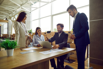 Young smiling business man showing work project to his colleagues on laptop computer while sitting at his workplace in the office. Smiling people chatting on a meeting discussing company finance.