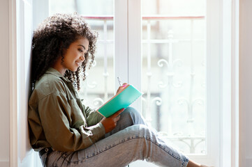 African woman with notebook writing sitting on windowsill at home