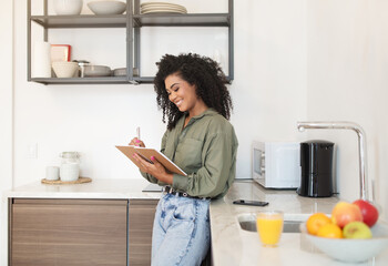 Happy brazilian woman reading and writing in notebook in kitchen