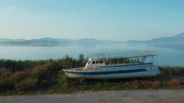Drone view of old abandoned pleasure or fishing boat on shore of lake or sea overlooking mountains. Rusty and rotten boat washed ashore at dawn in sunshine. Fishing village in Turkey.