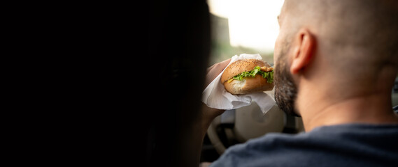 Millennial man eating a hamburger in a car while driving, view over his shoulder. Banner.