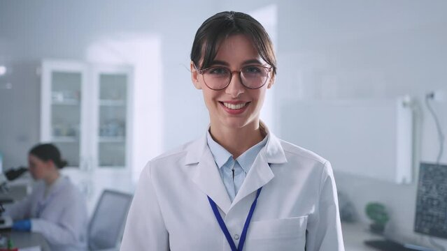Close-up portrait of good-looking female biologist in glasses and white lab coat looking at camera in modern laboratory room. Young Caucasian woman posing in workplace. Profession, job concept