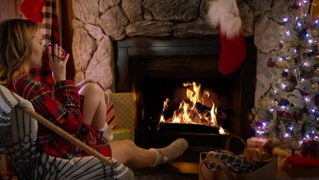 Woman relaxing by glowing burning fire and warming up by hot tea or hot chocolate. Winter and Christmas holidays concept, RED camera shot in slow motion. Female in woolen knitted socks at fireplace 4K