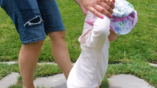 Close-up of cute baby girl walking barefoot with help of mother in summer park, slow motion