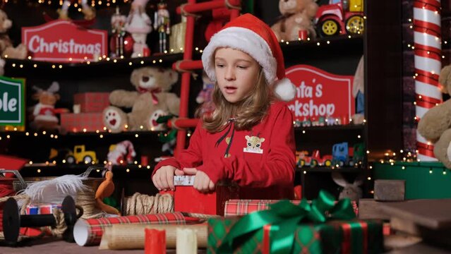 Cute Little Girl Santa helper Elf wrapping gifts at workshop, putting stuffed toy into a box at toy factory of Santa Claus