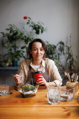 Asian florist looking at camera and holding coffee to go near terrarium and gardening tools in floral shop