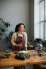 Smiling asian florist holding coffee to go near smartphone, notebook and succulents in floral shop