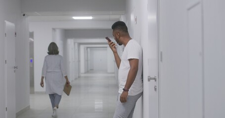 African American man stands in hospital corridor, records voice message and chats by mobile phone. Recovering patient during treatment in modern clinic. Medical staff walk in background. Slow motion.