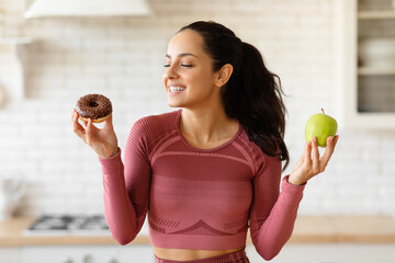 Athletic lady in sportswear choosing apple or donut in kitchen