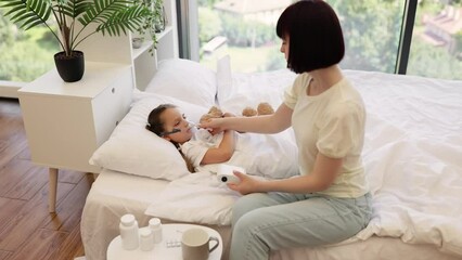 Brunette adult woman sitting on white bed and giving nebulizer inhaler to little ill daughter lying under blanket in bright room. Caucasian caring mom treating and helping kid recovering at cozy home.