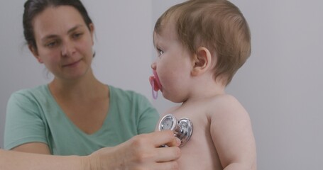 Handsome child sits at doctor checkup and puts pacifier in mouth. Pediatrician uses stethoscope to...