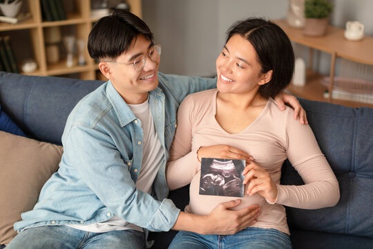 Korean couple expecting baby posing holding sonogram photo at home