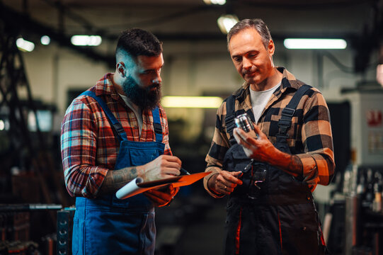 A quality specialist examining a metal part with his assistant taking notes.
