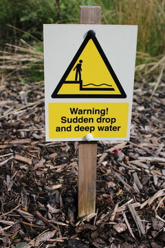 Yellow and white sign attached to a post placed in ground warning of sudden drop "Warning Sudden Drop and Deep Water"