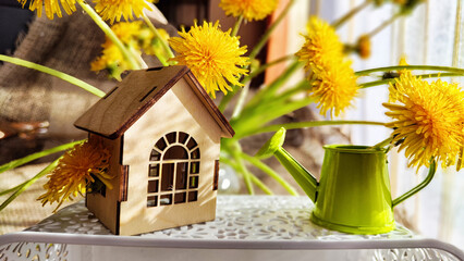 miniature toy house in dandelion flowers and watering can. natural background. symbol of family and...