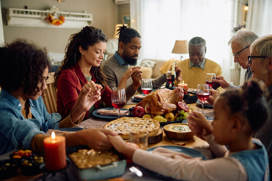 Multiracial extended family holding hands and praying while gathering for Thanksgiving meal at dining table.