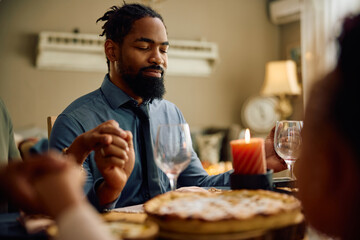 Black man praying with his family during Thanksgiving dinner at dining table.