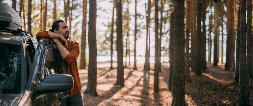 A young man stands next to a car with an upper tourist trunk in a pine forest on the shore of a lake at sunset.