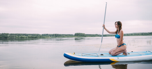 Naklejka premium A young woman with an open swimsuit swims on a SUP board on a picturesque lake. Evening tour. A beautiful, slender girl is engaged in sap surfing on the calm water of a picturesque pond.