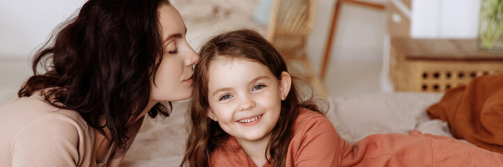 Beautiful young mother kissing happy cute little girl daughter lying on bed in bedroom. Mom and...