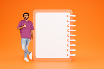 Smiling young indian guy in casual show thumb up with hands near list with free space, approve work...