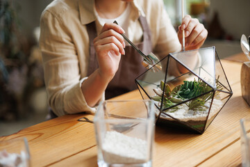 Partial view of craftswoman holding gardening tools while creating terrarium with succulents in...