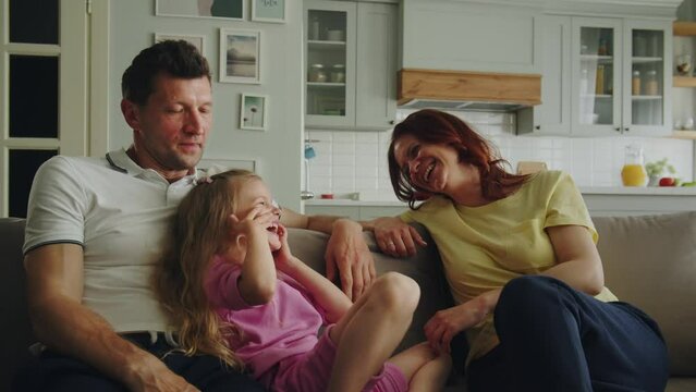 A happy american dream family is playing rock paper scissors in the room on the sofa. Mother won and laugh with daughter merrily. Father tickles her. Pleasant good nostalgia for children's games. 