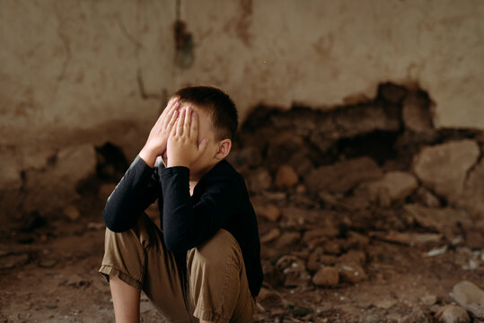 A sad little child, a preschooler, sits on the ruins of his destroyed house after the arrival of a rocket in dark clothes and cries
