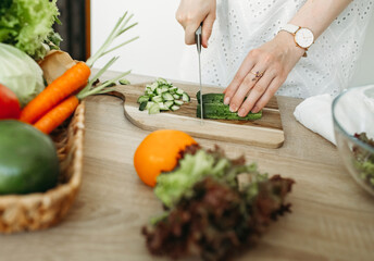 A woman is cutting a cucumber, vegetables lie next to her on the kitchen table.