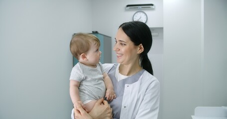 Female pediatrician stands in middle of bright hospital room and looks at camera. Adult doctors holds baby. Little boy looks at camera and holds hand of medic. Medical staff at work in modern clinic.