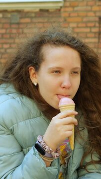 Teenager girl licks scoop of ice cream in waffle cone hesitantly and carefully. Female teen slightly touches ice cream with tongue on warm and sunny autumn day in urban area. Vertical video.
