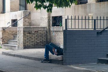 Homeless person lying on an old matrass, legs only visible, in the recess of a brick building with a beer can between his arm and body clearly sleeping or passed out