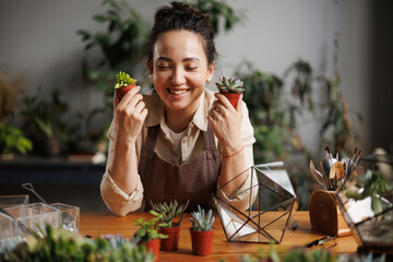 Cheerful asian florist in apron holding succulents near gardening tools and florariums in floral shop