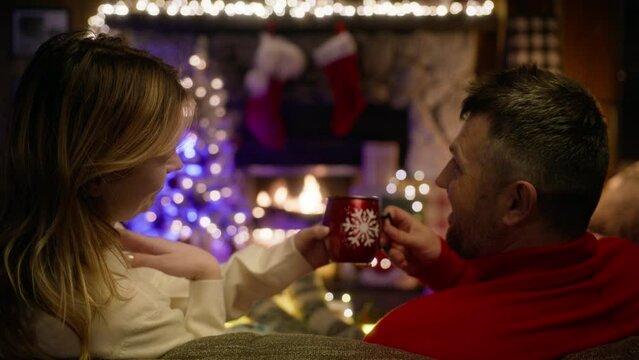 Boyfriend and girlfriend enjoying hot drink and conversation on Christmas holiday vacation closeup. Romantic couple embrace cuddle on winter vacation. Two lovers enjoying cozy fireplace back view 4k
