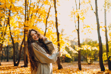 Smiling young woman plays and jumping dancing in the autumn park with the yellow leaves at sunset