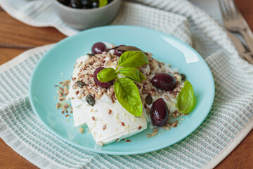 Feta cheese sliced on blue plate with sesame, flax seeds, served with black olives and basil leaves. Delicious and healthy breakfast on terrace