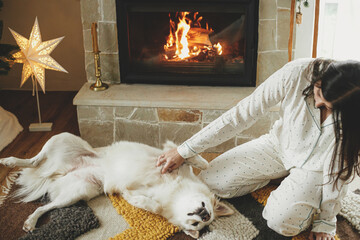Cozy christmas morning. Beautiful woman in stylish pajamas relaxing and playing with cute white dog...