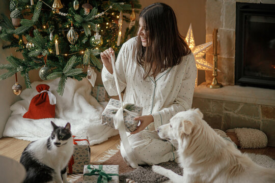 Cozy Christmas Morning. Beautiful Woman In Pajamas Opening Stylish Christmas Gift With Cute White Dog And Cat At Fireplace And Decorated Tree In Festive Living Room. Merry Christmas!