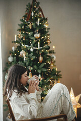 Beautiful woman in stylish pajamas with warm tea relaxing at decorated tree, enjoying cozy christmas morning in festive living room. Merry Christmas! Winter hygge