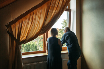 A happy elderly couple stand near the window, look.