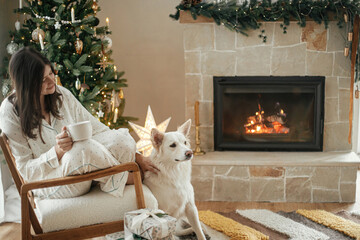Cozy christmas morning. Beautiful woman in stylish pajamas relaxing with warm tea and cute dog at...
