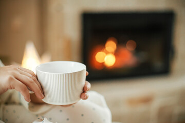 Cozy Christmas eve. Close up of warm cup of tea in hands on background of burning fireplace and glowing christmas star in festive living room. Merry Christmas and happy holidays!