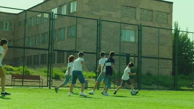 Group of boys playing football on  sport playground in sunny day . Many kids play soccer in stadium with green grass and gate . Training , competition , healthy activity for children . Slow motion