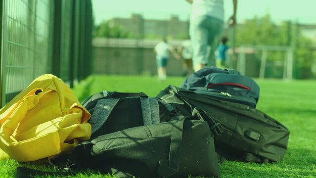 Kids throwing their backpacks on the grass ground and running to play football after school lessons . Close up many bags or schoolbags on the floor . Children playing soccer on background in sunny day