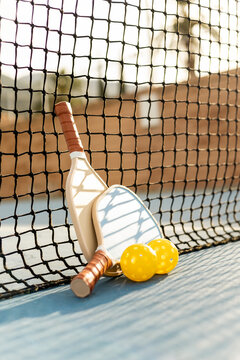 Photograph of two pickleball rackets and two balls next to a net on a sports court at sunset. Pickle ball concept. Sports similar to tennis.