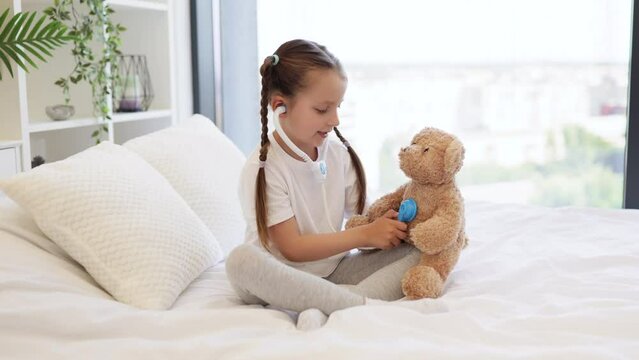 Adorable caucasian girl dressed in domestic clothes using toy stethoscope to checking respiratory system of small teddy bear. Cute doctor with braids hairstyles spending time with pleasure at home.