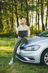 A cheerful man in glasses sits on the hood of his car in nature and works on a laptop. Freelance work, remote work during vacation