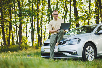 A successful man in glasses works on a laptop while leaning on the hood of his car. Remote work in nature. Work on a laptop online. Working on a computer while traveling.