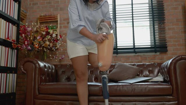 Asian disabled woman takes off her prosthetic leg to sit on a sofa using a laptop.
